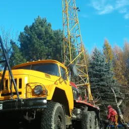 Żółty samochód ciężarowy z wysoką, żółtą wieżą wiertniczą na tle błękitnego nieba i drzew. Obok siedzi osoba w czerwonej kurtce na krześle.