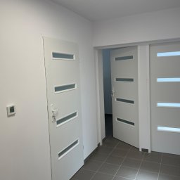 Firma Handlowo Usługowa - Wnętrze przedpokoju z trzema białymi drzwiami z poziomymi przeszkleniami, szara podłoga z płytek, termostat na ścianie.