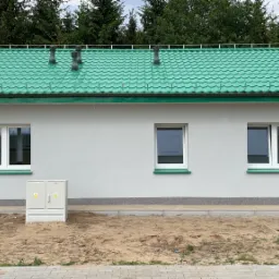 Nowo otynkowany budynek z zielonym dachem z blachy trapezowej, widoczne trzy okna z białymi ramami, szara elewacja i skrzynka elektryczna przy ścianie