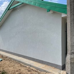 Firma Handlowo Usługowa - Świeżo otynkowana, jasnoszara elewacja domu jednorodzinnego z zielonym dachem, widoczny cokół z szarego kamienia i fragment chodnika z kostki brukowej.