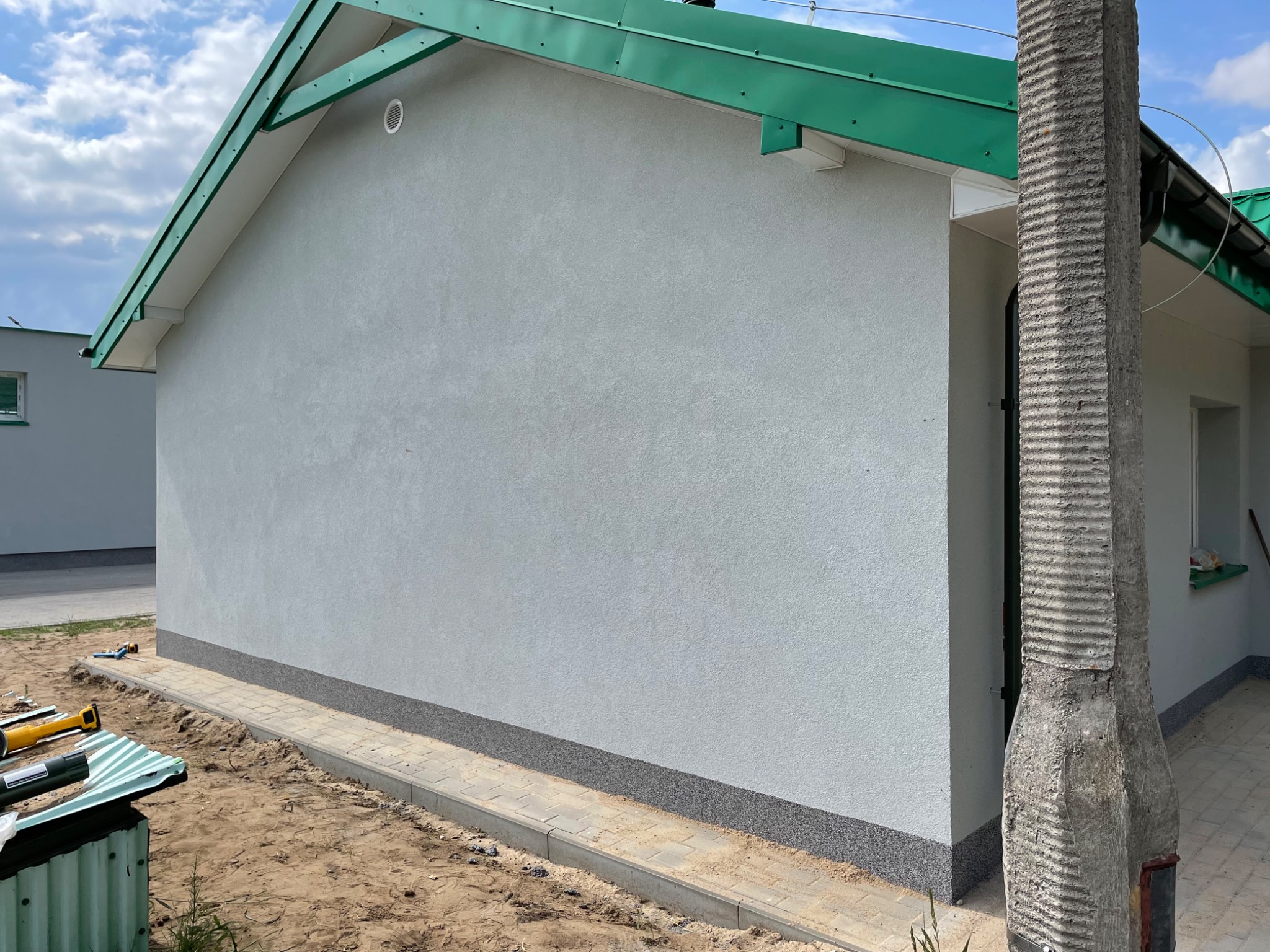 Świeżo otynkowana, jasnoszara elewacja domu jednorodzinnego z zielonym dachem, widoczny cokół z szarego kamienia i fragment chodnika z kostki brukowej.