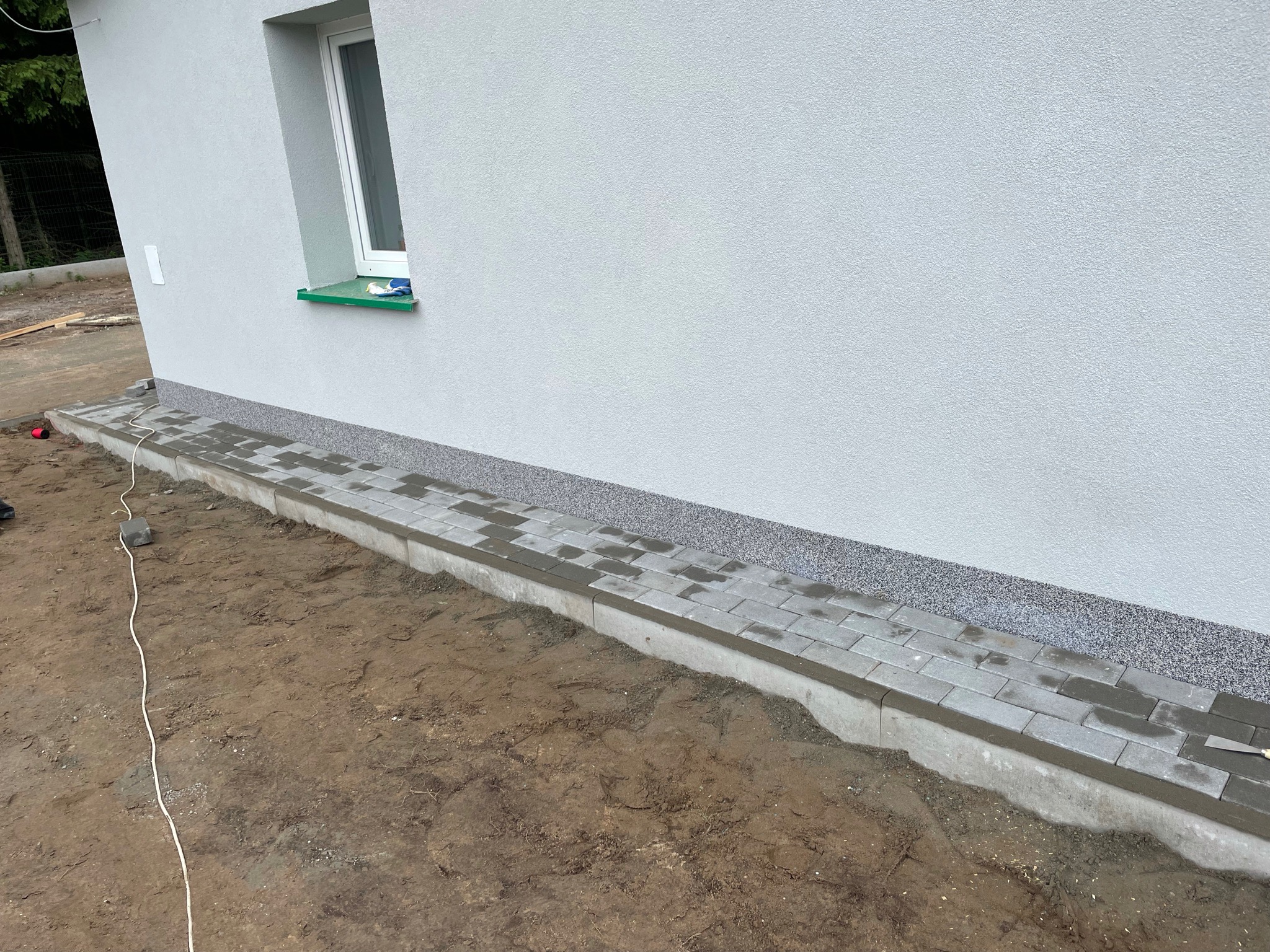 Świeżo ułożona kostka brukowa przy elewacji domu, widoczny fragment ściany z oknem i obrzeże z szarego grysu.