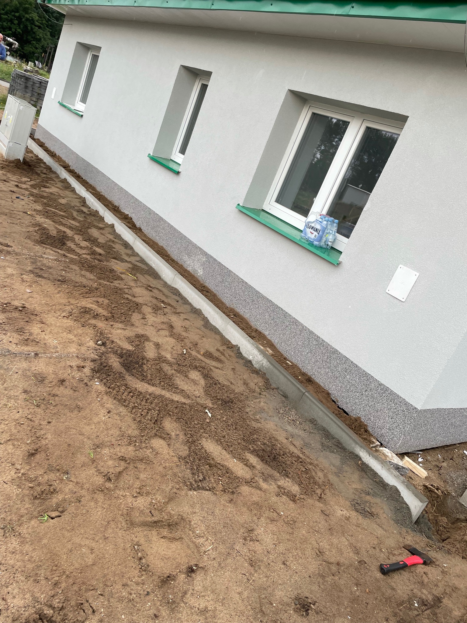Świeżo otynkowana elewacja domu jednorodzinnego z widocznymi oknami z zielonymi parapetami, ułożonym obrzeżem chodnikowym i ziemią dookoła. Na parapecie butelki z wodą.