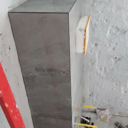 Pionowy element obudowany szarymi płytkami imitującymi kamień, z widocznymi akcesoriami do układania płytek, poziomica i surowa ściana w tle.