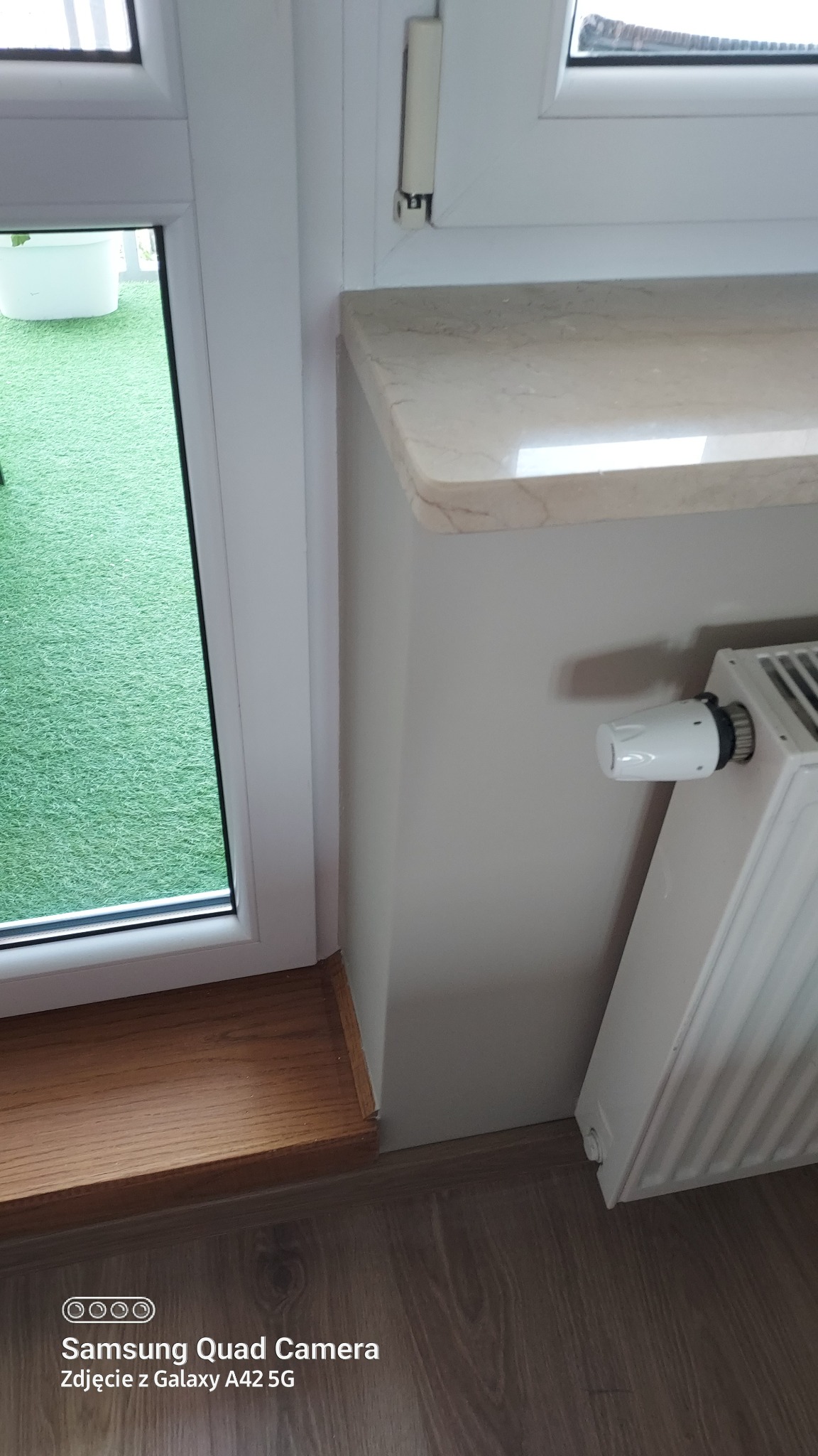 Fragment wnętrza z oknem, marmurowym parapetem, grzejnikiem i drewnianą podłogą, widok na zielony trawnik za oknem.