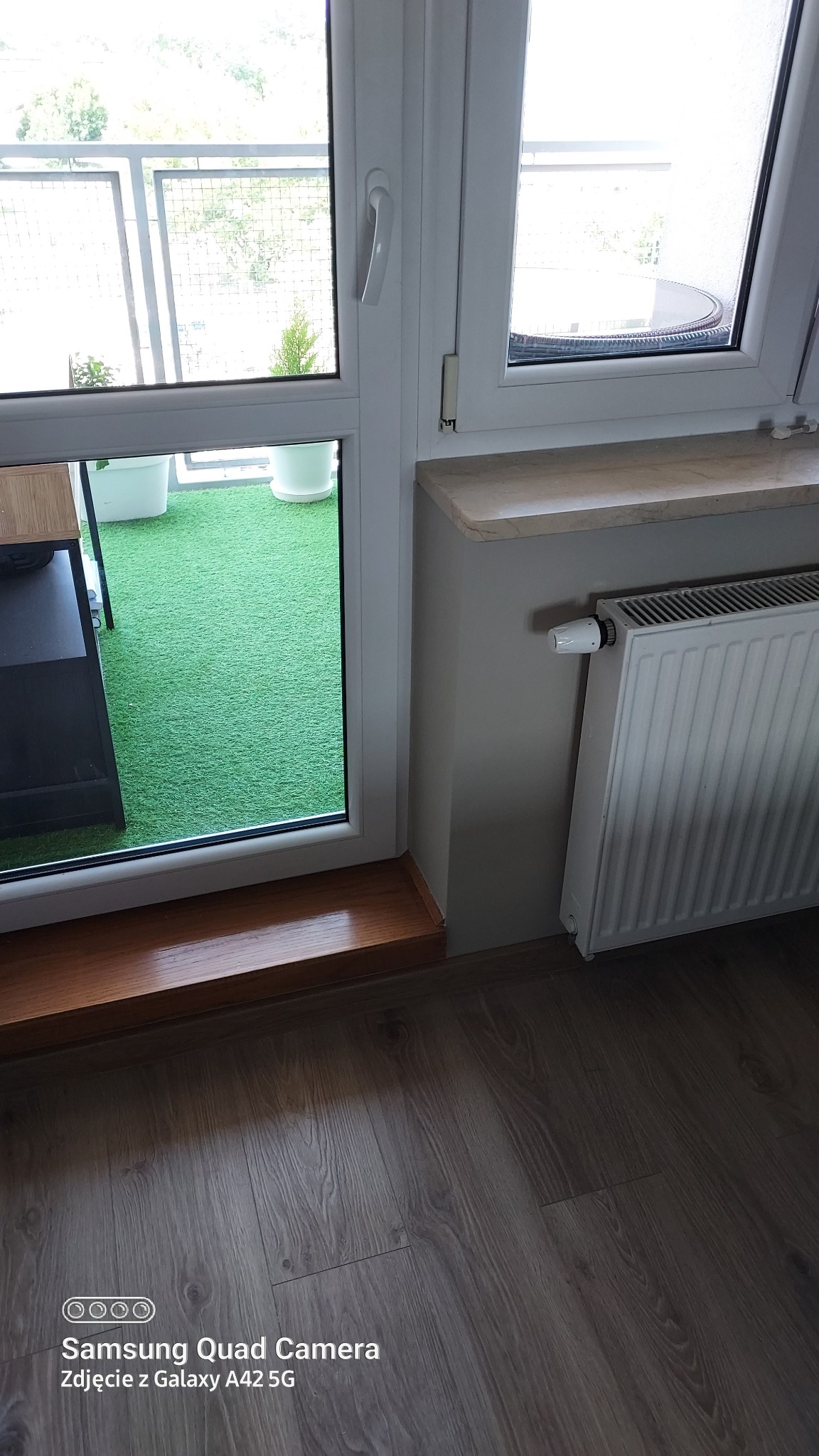 Fragment pokoju z widokiem na balkon z zieloną sztuczną trawą, widoczne panele podłogowe, białe okno balkonowe z drewnianym parapetem wewnętrznym i grzejnik.