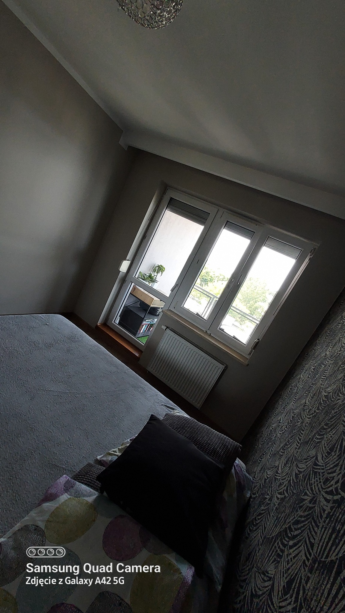 Pokój po remoncie, widok na świeżo pomalowane ściany w odcieniach szarości, okno z widokiem na zewnątrz, grzejnik pod oknem, łóżko z poduszkami w tle, wzorzysta tapeta na ścianie.