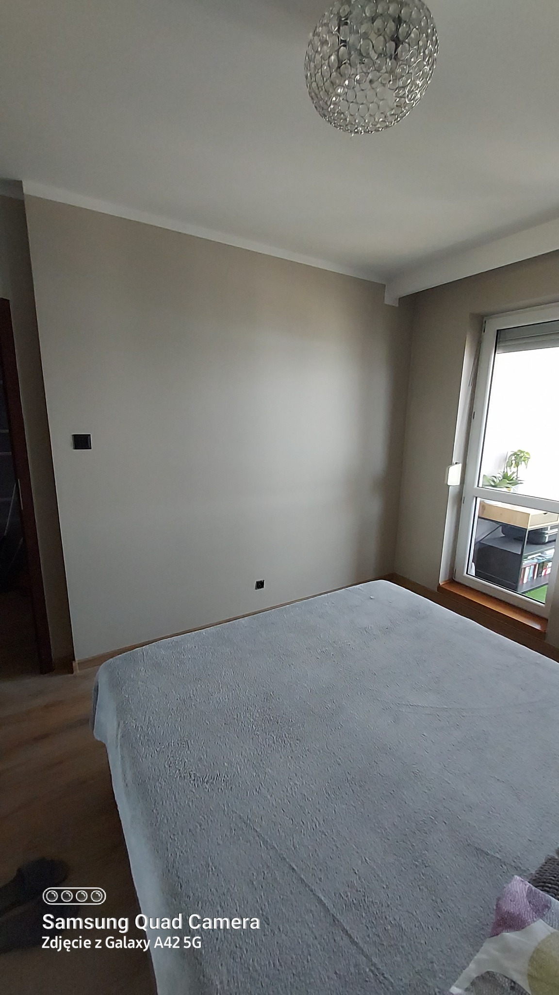 Pokój w mieszkaniu we Włocławku po malowaniu na beżowo, widoczny sufit z ozdobną lampą, ściany z gładzią oraz fragment łóżka z szarą narzutą.