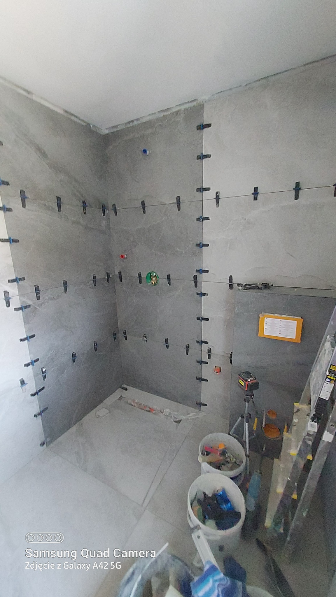 Świeżo ułożone, szare płytki w łazience, zabezpieczone krzyżykami dystansowymi i żyłką, widoczny laser krzyżowy na statywie, dwa wiadra z narzędziami i drabina oparta o ścianę.