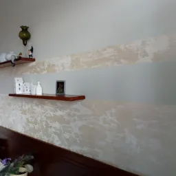 Ściana w sypialni z dekoracyjnym efektem malowania w odcieniach beżu i bieli, dwie półki z ozdobami, widoczny fragment wezgłowia łóżka i kwiat doniczkowy.