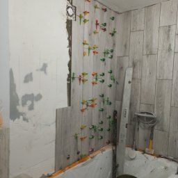 Tom-Serwis Tomasz Borucki - Układanie jasnych płytek ceramicznych imitujących drewno na ścianie łazienki, z widocznymi kolorowymi klipsami poziomującymi i zabezpieczoną wanną.