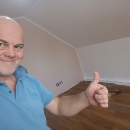 Tom-Serwis Tomasz Borucki - Zadowolony mężczyzna w niebieskiej koszulce pokazuje kciuk w górę, w tle układanie paneli laminowanych na podłodze w pokoju ze skośnym sufitem i dwukolorowymi ścianami.