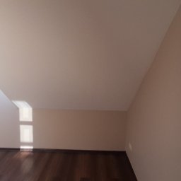 Tom-Serwis Tomasz Borucki - Poddasze z oknem dachowym, świeżo pomalowane ściany w odcieniach beżu i brązu, drewniana podłoga, widoczny fragment okna z widokiem na zewnątrz.