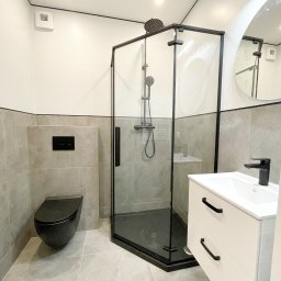 Projektowanie wnętrz Art Centrum Nieruchomości - Nowoczesna łazienka z czarnymi akcentami: kabina prysznicowa z czarną ramą, wisząca toaleta w kolorze czarnym, biała szafka pod umywalkę z czarnymi uchwytami, okrągłe lustro i szare płytki.