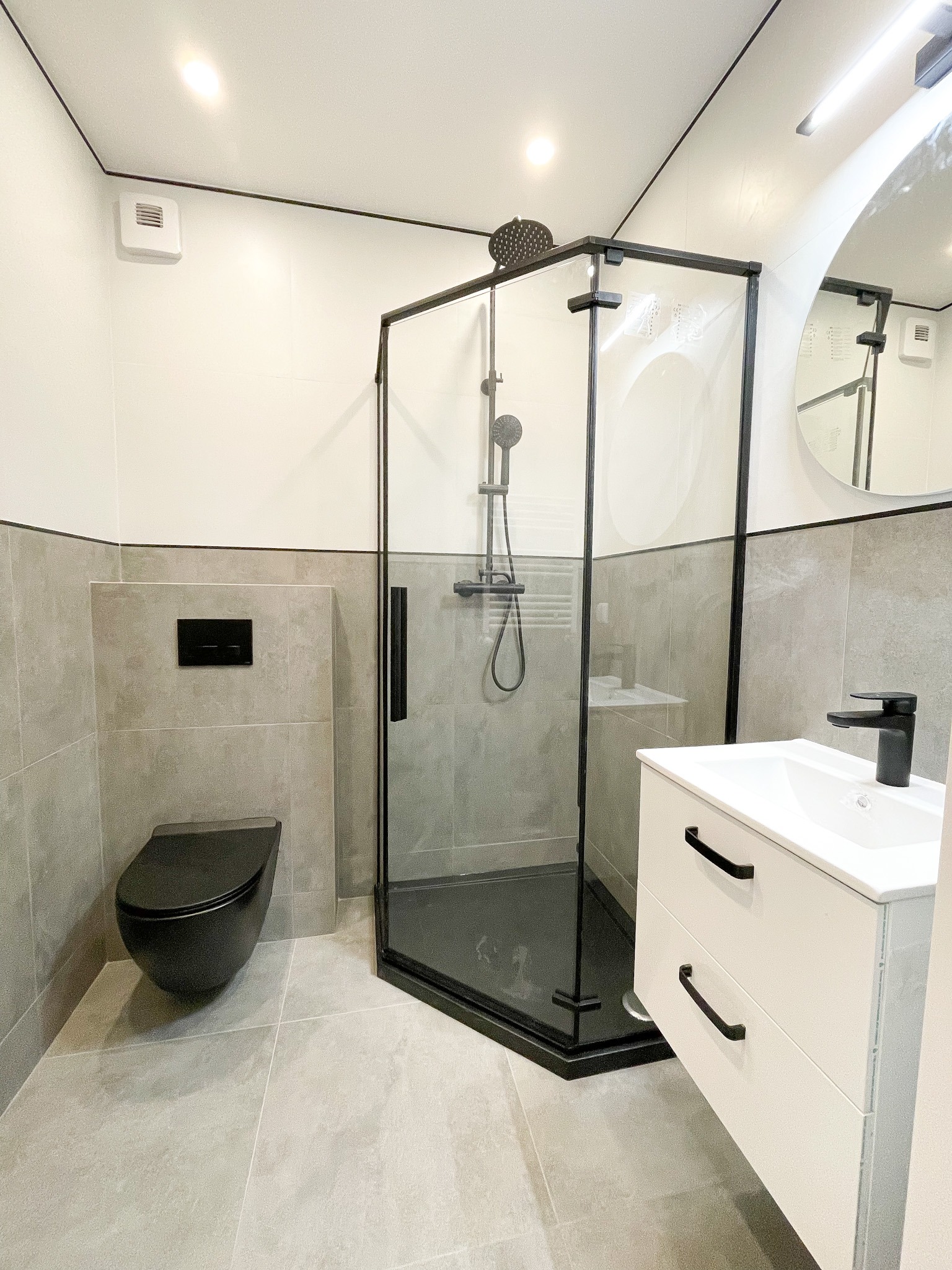Nowoczesna łazienka z czarnymi akcentami: kabina prysznicowa z czarną ramą, wisząca toaleta w kolorze czarnym, biała szafka pod umywalkę z czarnymi uchwytami, okrągłe lustro i szare płytki.