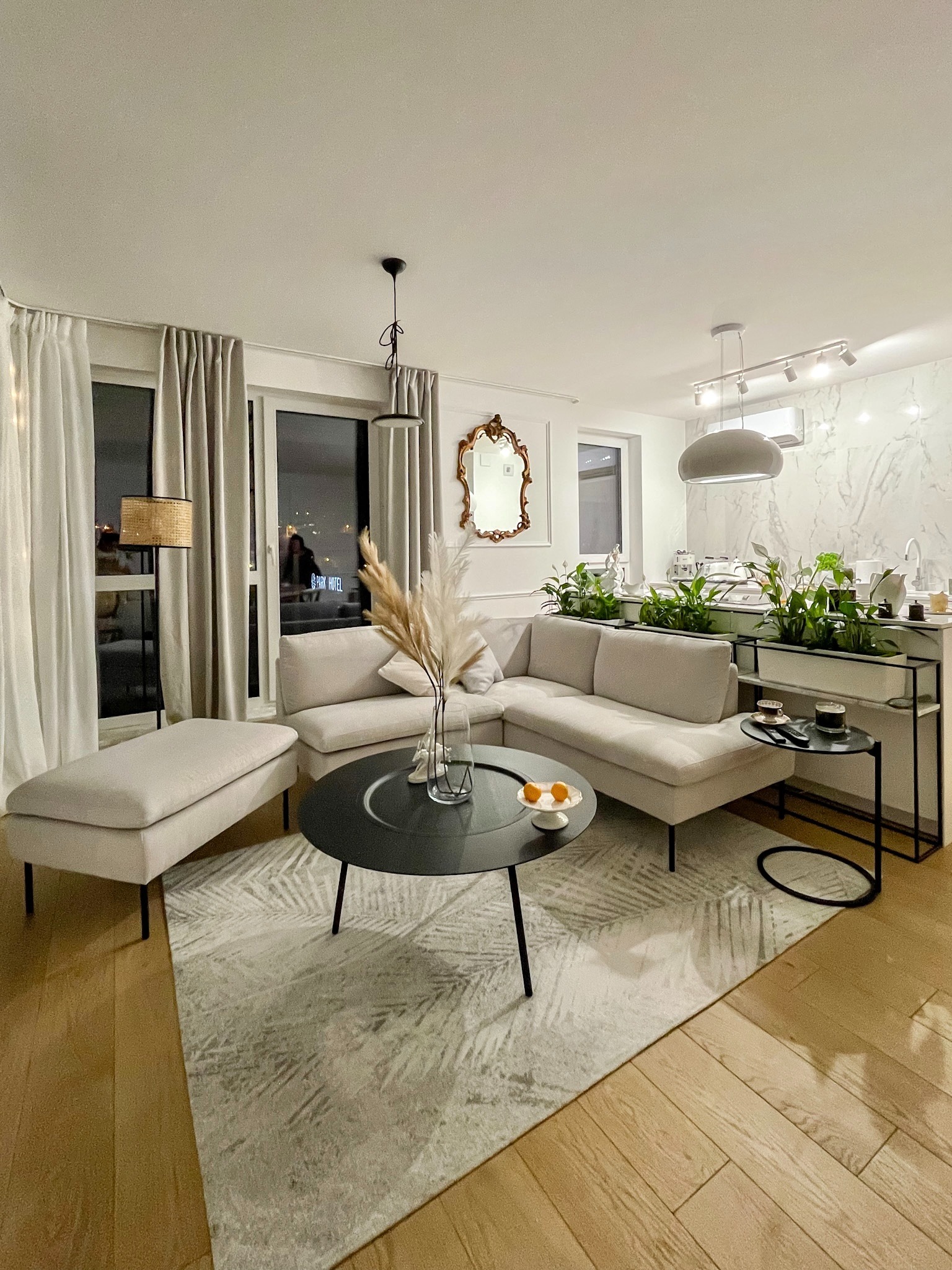 Jasny narożnik z pufem w salonie, okrągły czarny stolik kawowy z owocami, dywan w liście, parkiet, za sofą rośliny doniczkowe na blacie, widok na miasto nocą.