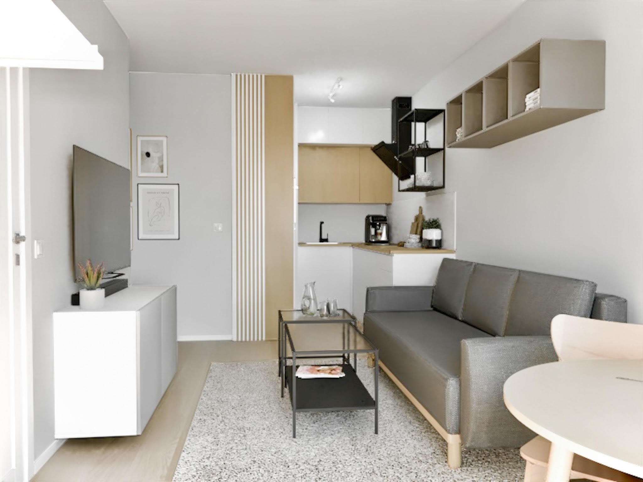 Jasne wnętrze mieszkania z szarą sofą, szklanym stolikiem kawowym, białymi szafkami RTV, elementami dekoracyjnymi w kolorze beżowym i otwartą kuchnią w tle.