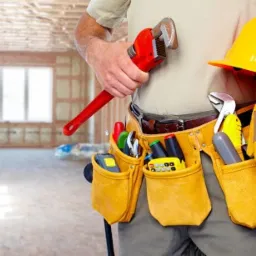 Mężczyzna z pasem narzędzi, kluczem nastawnym i kaskiem na tle surowego wnętrza w trakcie remontu.