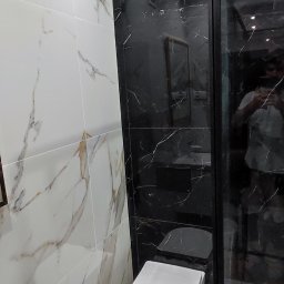 GlazuraProArt - Nowoczesna łazienka z białymi i czarnymi płytkami imitującymi marmur, wisząca toaleta, umywalka z szafką i czarna kabina prysznicowa z widocznym odbiciem osoby.