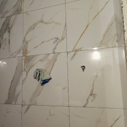 GlazuraProArt Oleksandr Kopyshchyk Prace wykończeniowe i remontowe w budownictwie - Ściana w trakcie układania płytek imitujących marmur w Legnicy, widoczna szmatka do czyszczenia i taśma maskująca przy suficie.