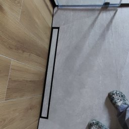 GlazuraProArt - Nowoczesna łazienka z odpływem liniowym, drewnopodobne płytki na ścianie, szare płytki podłogowe, kabina prysznicowa ze szkła i czarnych profili oraz osoba w klapkach moro.