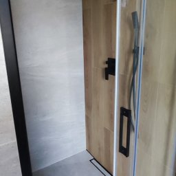 GlazuraProArt Oleksandr Kopyshchyk Prace wykończeniowe i remontowe w budownictwie - Nowoczesny prysznic walk-in z czarną armaturą, drewnopodobnymi płytkami na ścianie i szklaną kabiną.