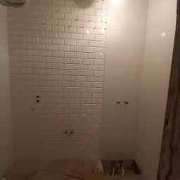 GlazuraProArt - Łazienka w trakcie prac wykończeniowych: białe, prostokątne płytki ułożone w cegiełkę na ścianie, widoczne instalacje hydrauliczne, sufit w trakcie montażu, podłoga zabezpieczona kartonem.