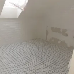 Biała łazienka na poddaszu w trakcie wykańczania, z ułożoną podłogą w geometryczny wz&oacute;r, białe płytki typu metro na ścianach i oknem dachowym z folią ochronną.