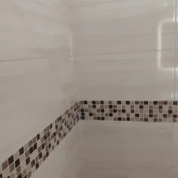 Fragment wykończonej łazienki z beżowymi, poziomymi płytkami imitującymi kamień, ozdobionymi pasem mozaiki z brązowych i beżowych kwadracików, widoczne połączenie ściany i podłogi z drewnopodobną...