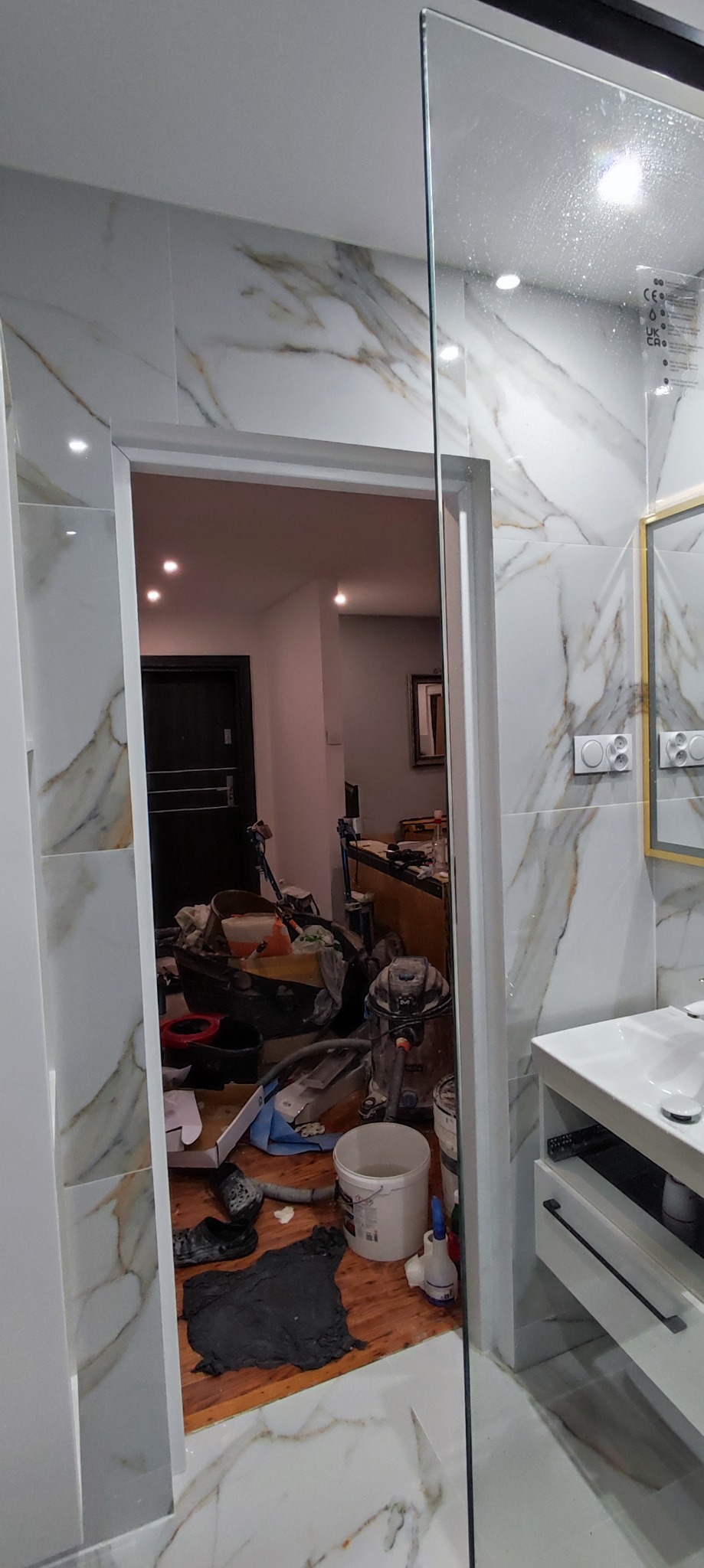 Łazienka w trakcie remontu z widocznymi narzędziami i materiałami budowlanymi, płytki imitujące marmur na ścianach, widok przez szklaną kabinę prysznicową na wnętrze z bałaganem.