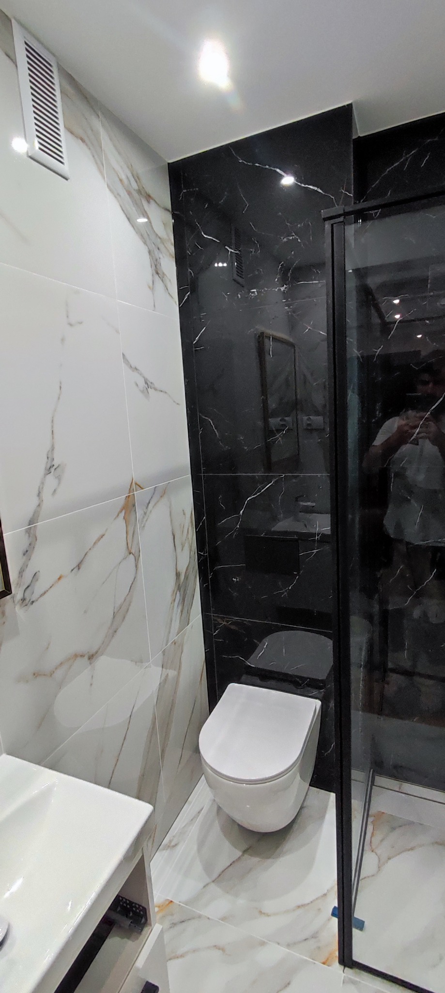 Nowoczesna łazienka z białymi i czarnymi płytkami imitującymi marmur, wisząca toaleta, umywalka z szafką i czarna kabina prysznicowa z widocznym odbiciem osoby.