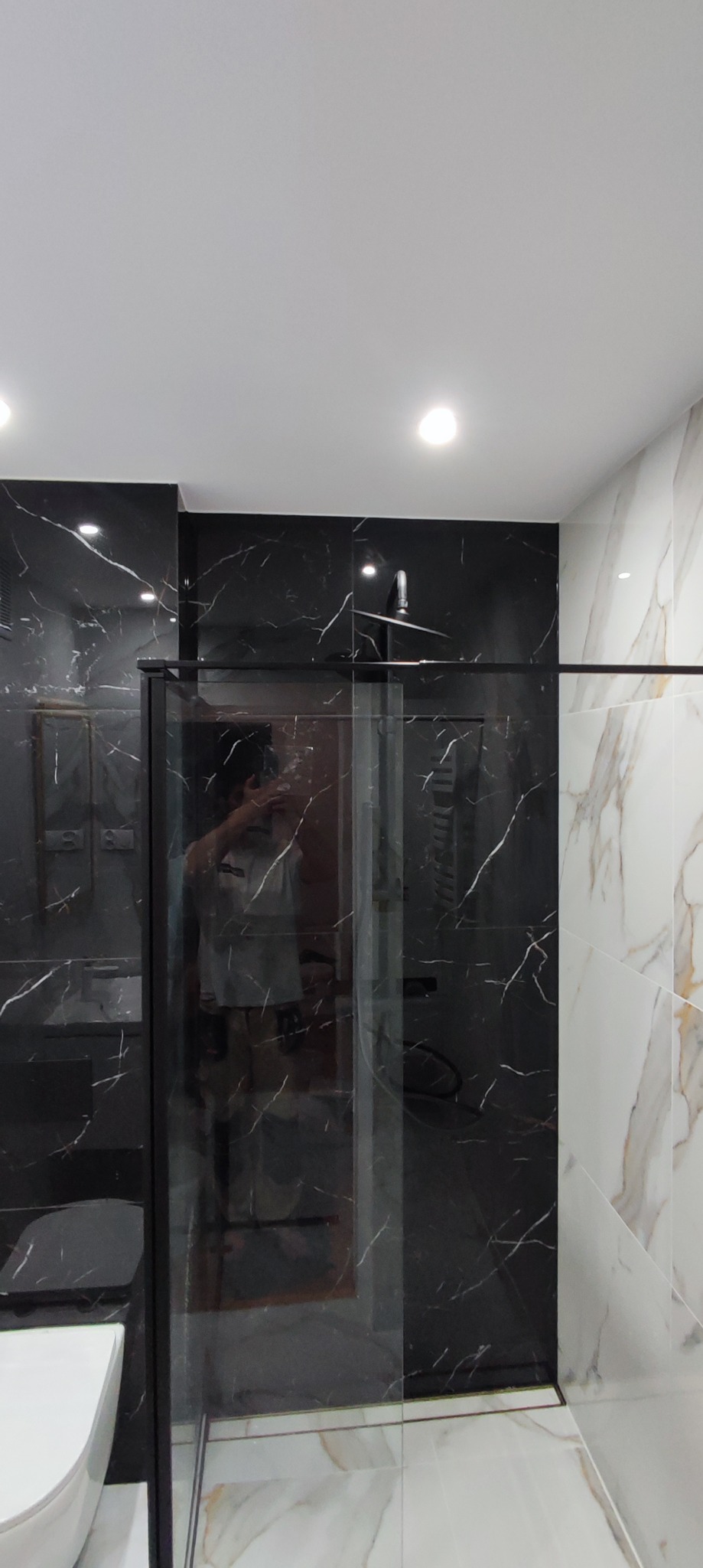Nowoczesna łazienka z kabiną prysznicową typu walk-in, wykończona płytkami imitującymi marmur w kolorze czarnym i białym, z czarną armaturą i oświetleniem punktowym.