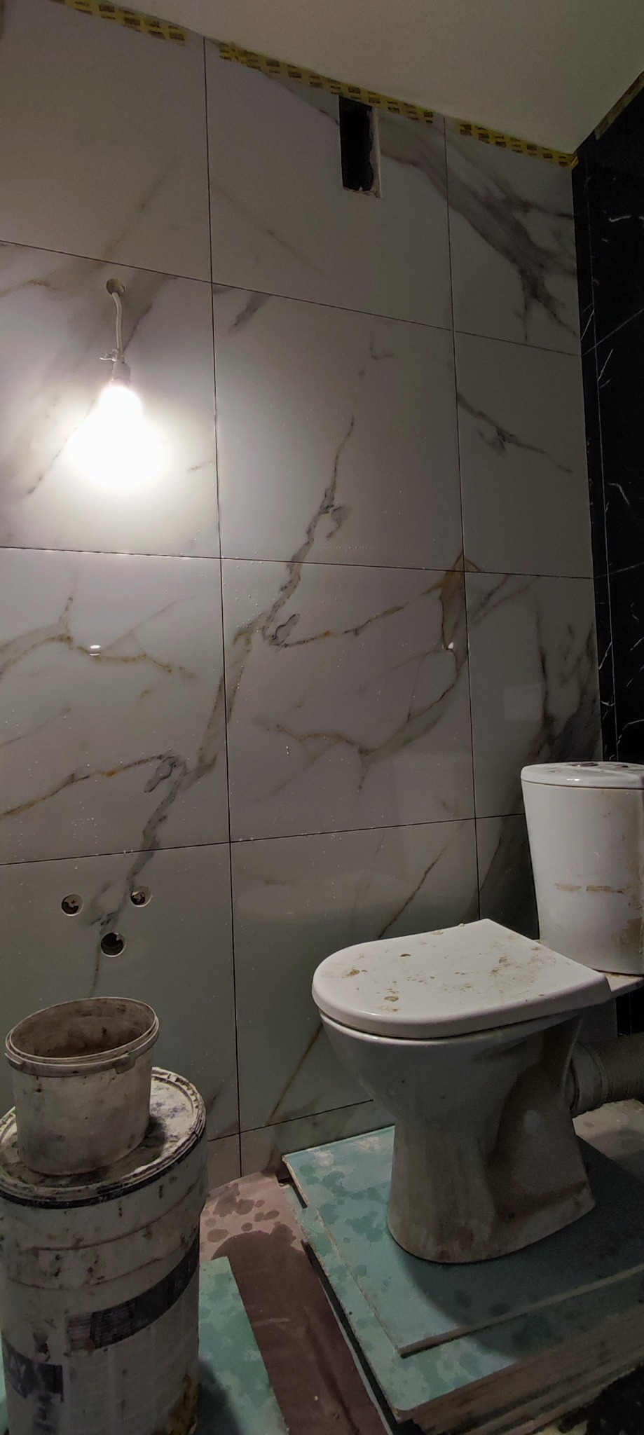 Białe płytki imitujące marmur na ścianie łazienki w trakcie remontu, widoczne otwory na instalacje, zaklejone taśmą zabezpieczającą górne krawędzie płytek, brudna toaleta zabezpieczona płytkami...