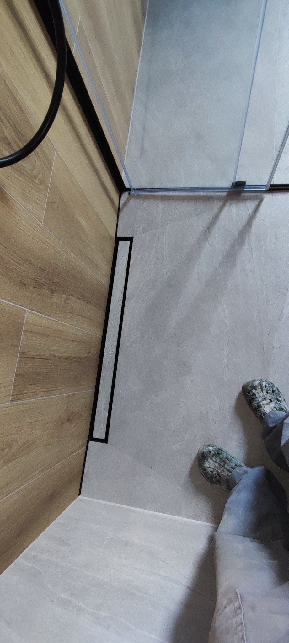 Nowoczesna łazienka z odpływem liniowym, drewnopodobne płytki na ścianie, szare płytki podłogowe, kabina prysznicowa ze szkła i czarnych profili oraz osoba w klapkach moro.