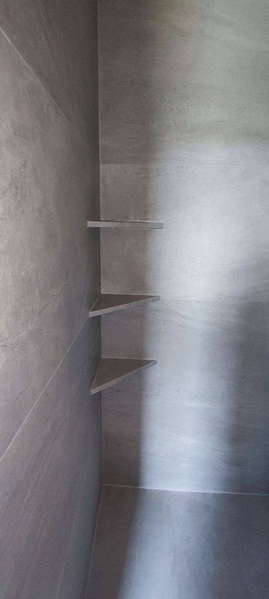 Szara, nowoczesna kabina prysznicowa wyłożona płytkami z trzema półkami narożnymi, bez widocznych akcesoriów.