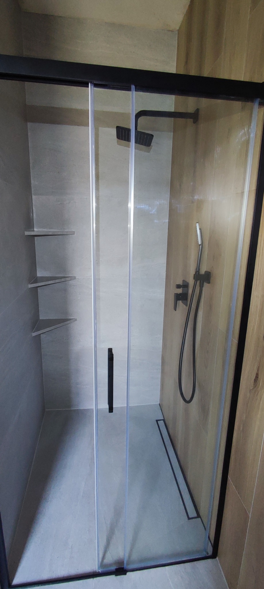 Nowoczesna kabina prysznicowa z czarnymi elementami, szklane drzwi przesuwne, szara płytka na ścianach i podłodze, drewnopodobna okładzina na jednej ze ścian, narożne półki wewnątrz.