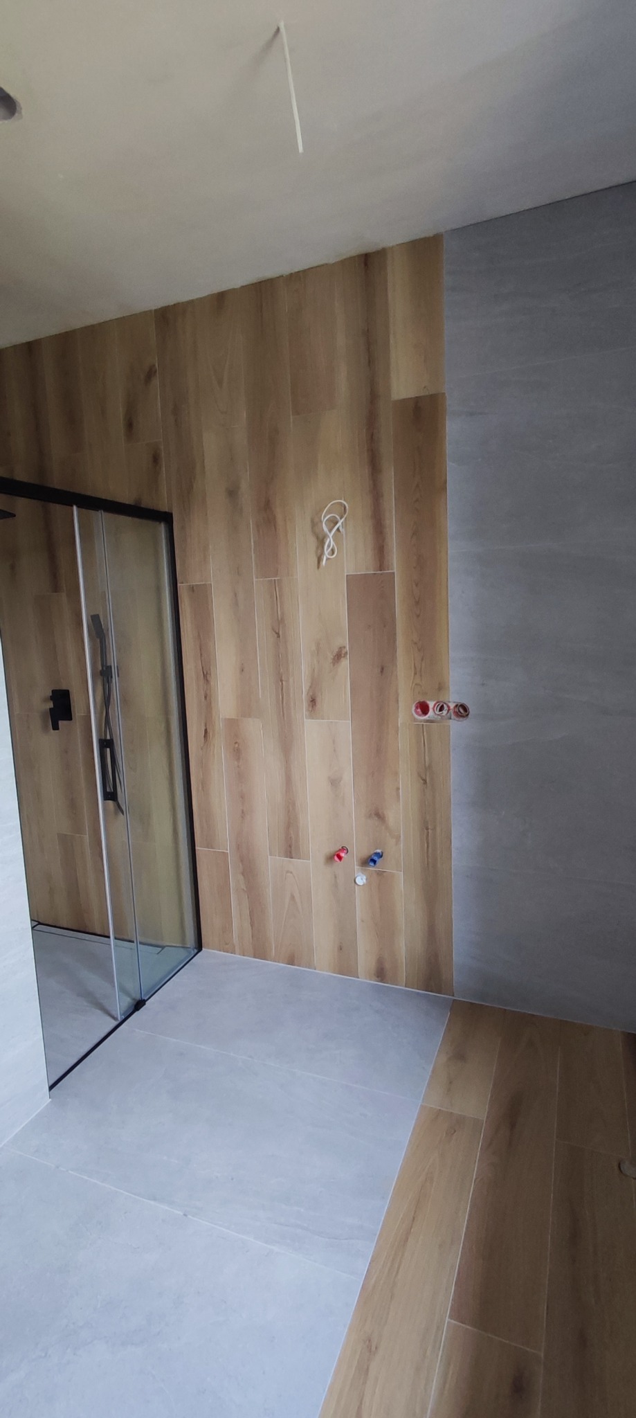 Wnętrze łazienki w trakcie wykańczania: kabina prysznicowa z czarnym wykończeniem, ściany wyłożone płytkami imitującymi drewno oraz szare płytki, widoczne wystające przewody i rury instalacyjne.