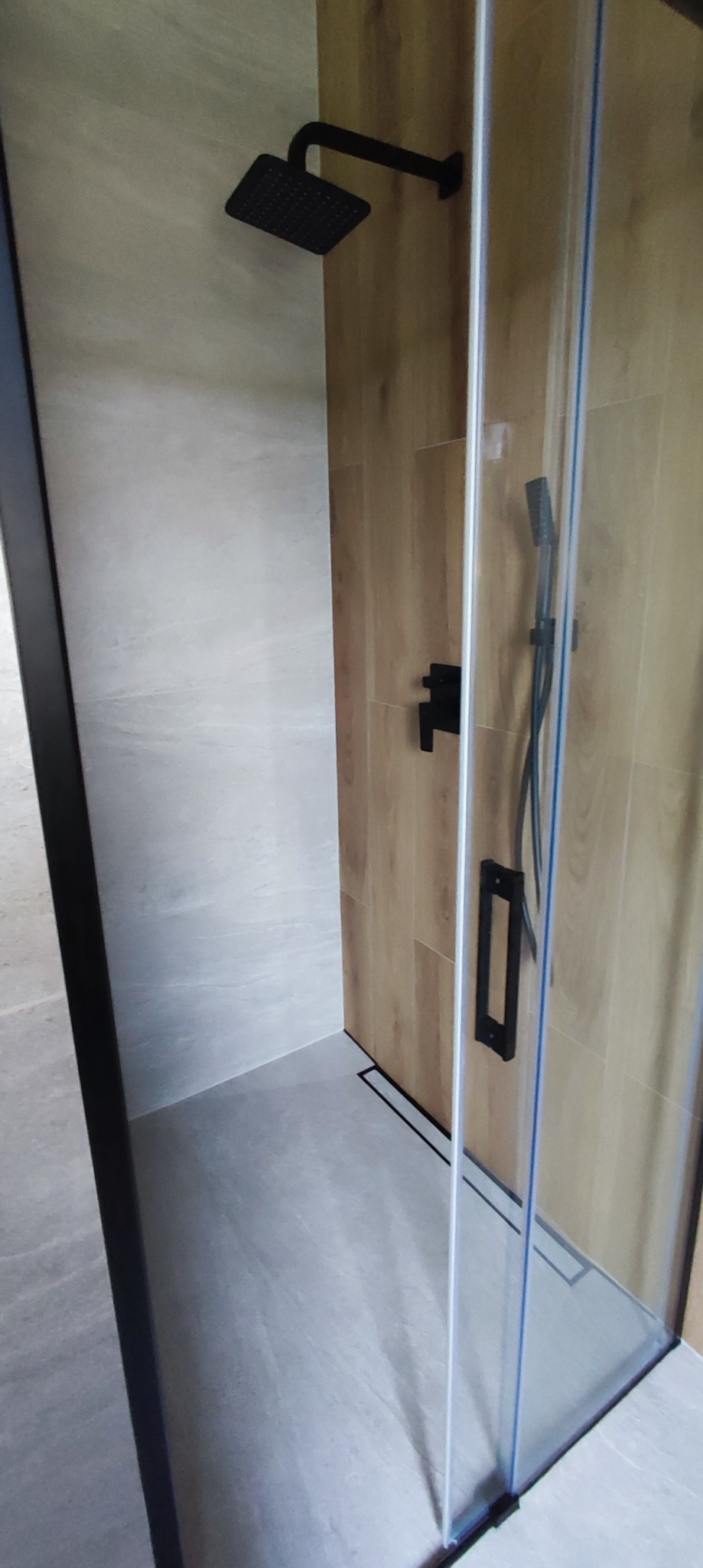 Nowoczesny prysznic walk-in z czarną armaturą, drewnopodobnymi płytkami na ścianie i szklaną kabiną.