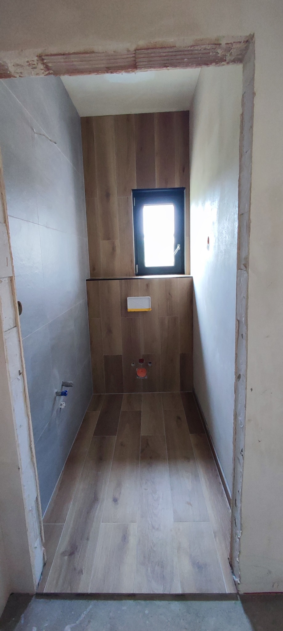 Wnętrze łazienki w trakcie remontu, widoczne płytki imitujące drewno na podłodze i ścianie, surowy stan ścian przy wejściu, okno z czarną ramą i częściowo zamontowany stelaż podtynkowy WC.