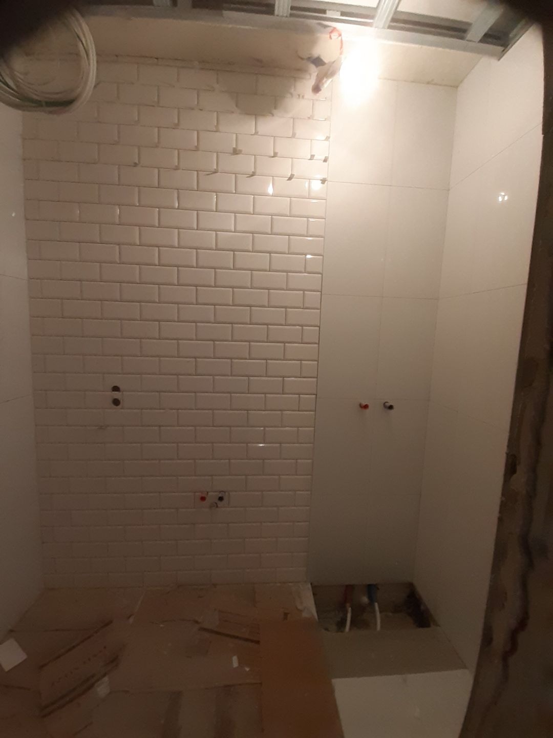 Łazienka w trakcie prac wykończeniowych: białe, prostokątne płytki ułożone w cegiełkę na ścianie, widoczne instalacje hydrauliczne, sufit w trakcie montażu, podłoga zabezpieczona kartonem.