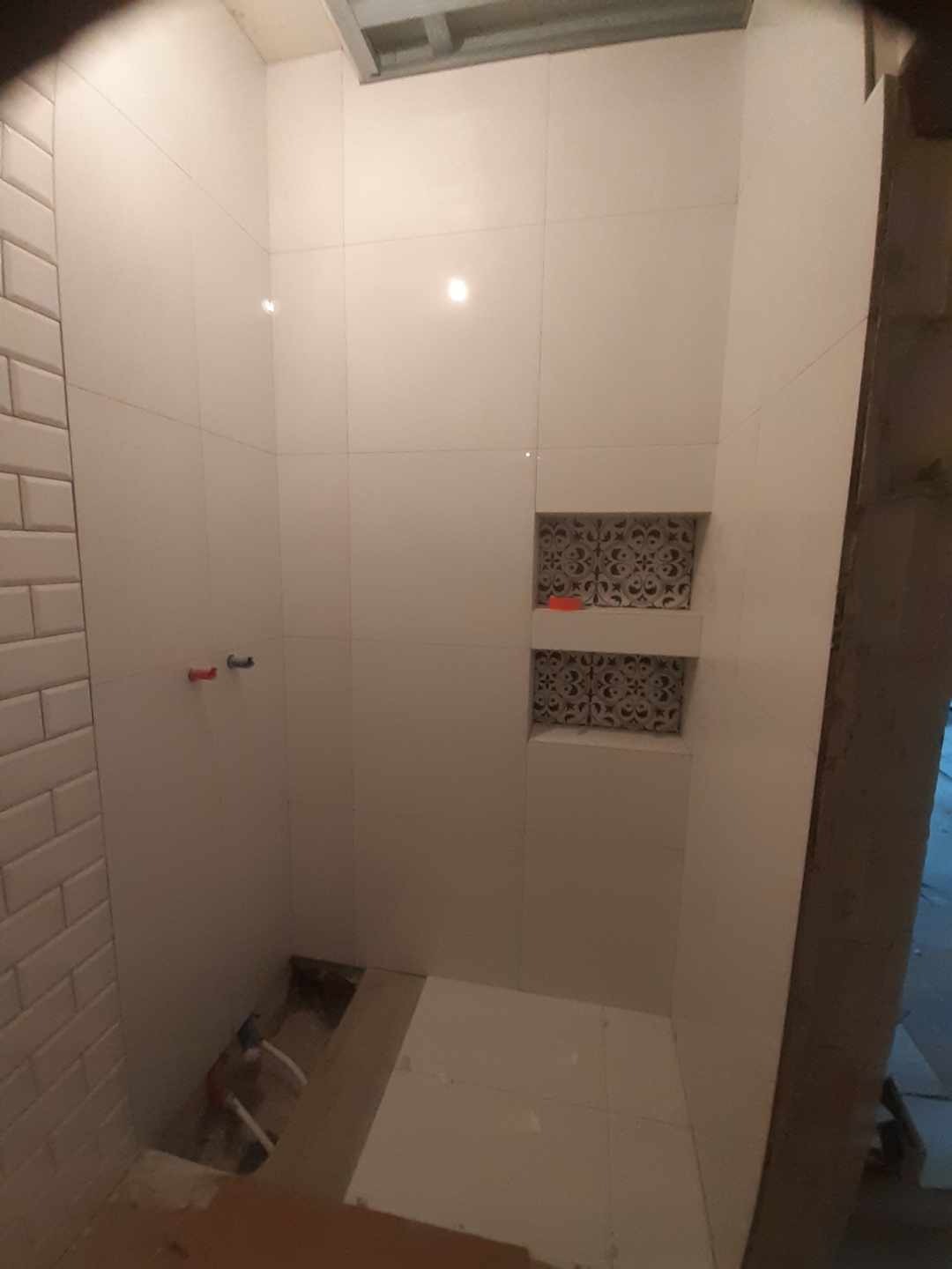 Wnętrze łazienki w trakcie prac wykończeniowych: białe płytki ścienne i podłogowe, wnęki dekoracyjne z wzorzystymi płytkami, widoczne rury instalacyjne i fragment sufitu podwieszanego.