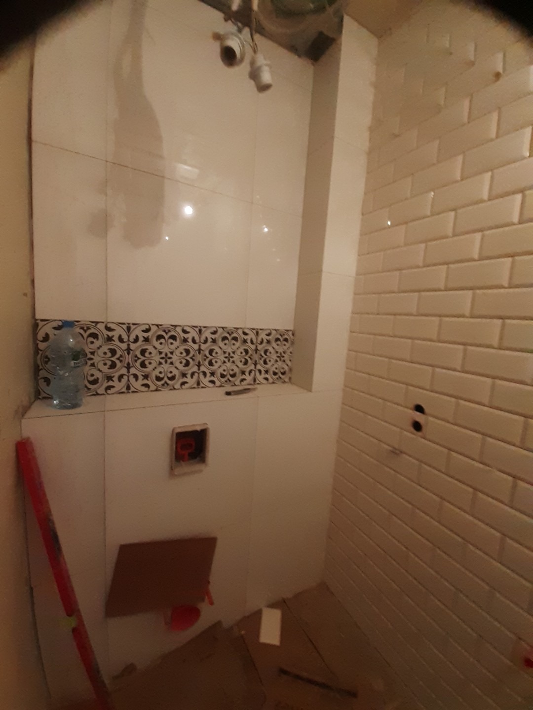 Remont łazienki w trakcie: białe płytki ścienne, dekoracyjny pas z wzorem, przygotowane otwory na instalacje, widoczny poziomica i butelka wody