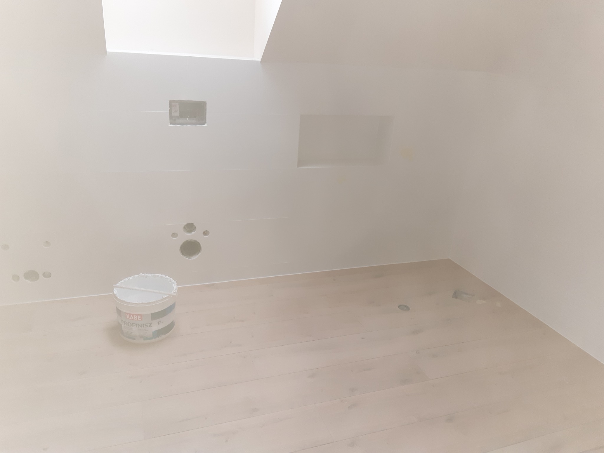 Białe, surowe wnętrze poddasza z drewnianą podłogą, widoczne otwory w ścianie i wiadro z białą farbą Kabe Profinisz, przygotowane do malowania.