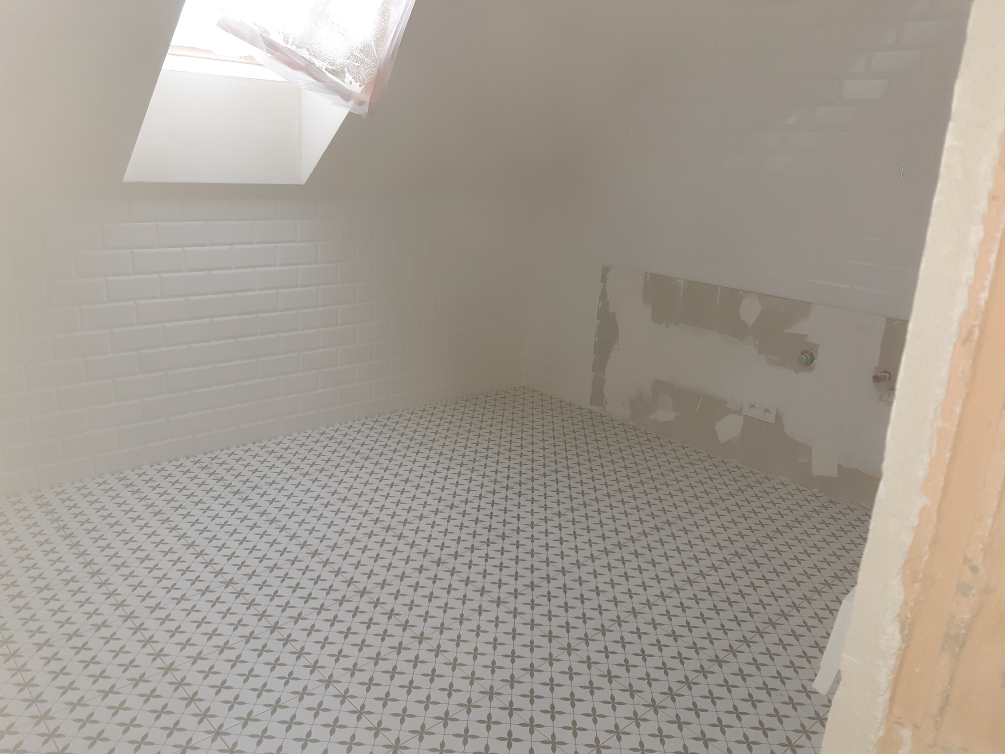 Biała łazienka na poddaszu w trakcie wykańczania, z ułożoną podłogą w geometryczny wzór, białe płytki typu metro na ścianach i oknem dachowym z folią ochronną.