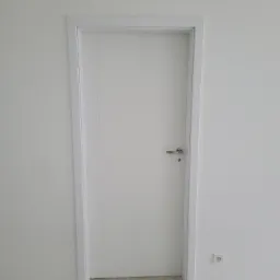 Nowoczesne, białe drzwi wewnętrzne z srebrną klamką, osadzone w białej ościeżnicy, na tle białej ściany i podłogi z paneli imitujących jasne drewno; widoczne gniazdko elektryczne przy podłodze.