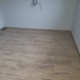 Ułożona podłoga z paneli imitujących drewno w nowym, jasnym pomieszczeniu z szarymi ścianami, widoczne kable wystające ze ściany i narzędzia w rogu.