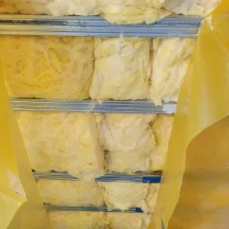 Ocieplenie poddasza wełną mineralną, widoczne metalowe profile i folia paroizolacyjna, fragment sylwetki osoby w zielonej kurtce.