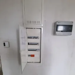 Otwarta skrzynka rozdzielcza z bezpiecznikami na ścianie w surowym stanie, obok zamknięta skrzynka z wentylacją i mały panel sterowania.