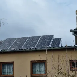 Panele słoneczne zamontowane na dachu domu jednorodzinnego, widoczne okna z firankami, rynna i antena na dachu, pochmurne niebo w tle.