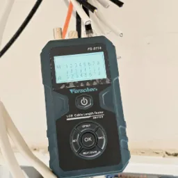 Miernik długości kabli Forscher FS-8114 z wyświetlaczem LCD, podłączony do przewodów elektrycznych w rozdzielnicy, widoczne bezpieczniki i złącza.